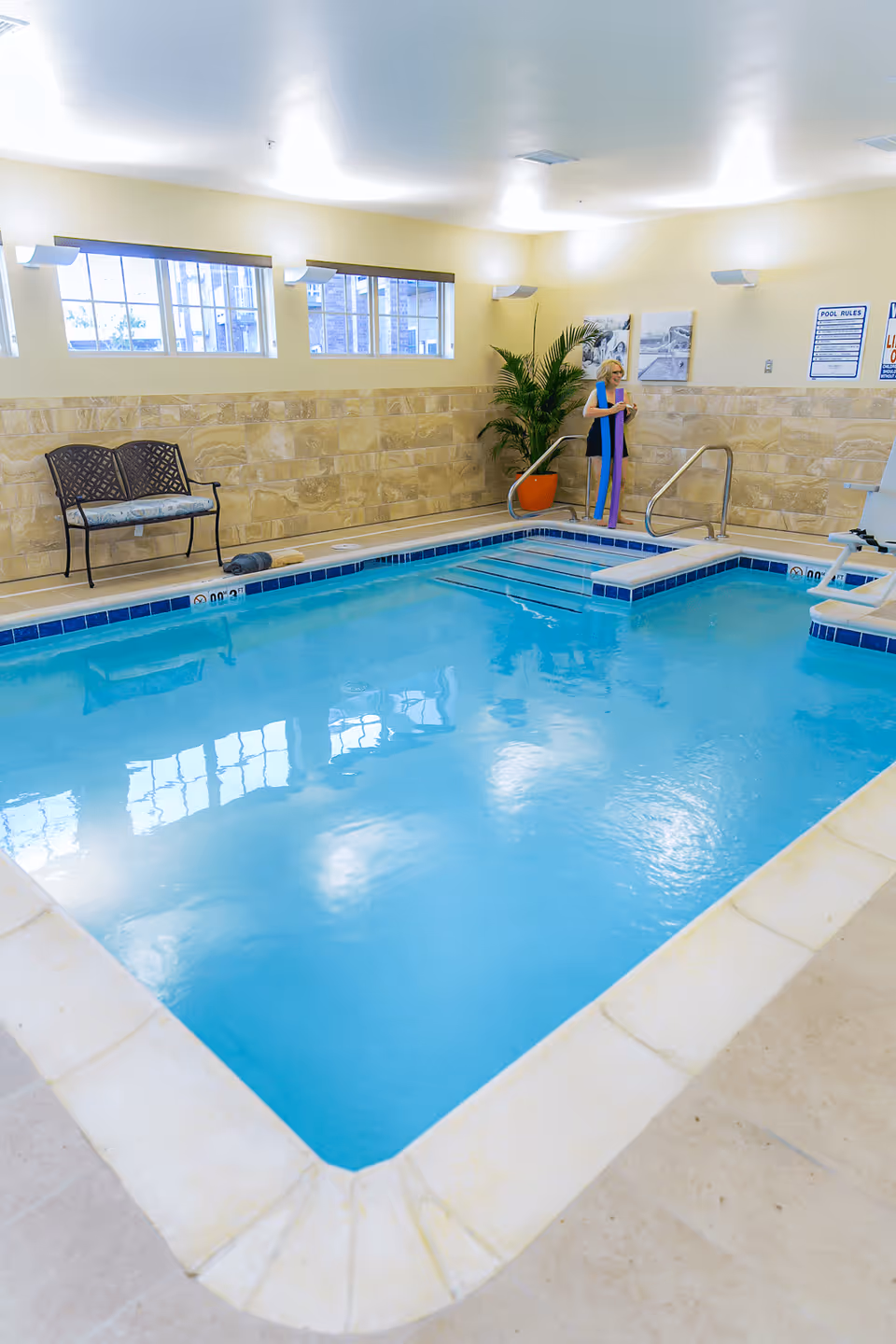 Indoor swimming pool area with clear blue water, beige tiled floor, and stone-textured walls. A woman stands near the pool steps holding a drink, next to a potted plant. There is a bench along the wall and windows letting in natural light.