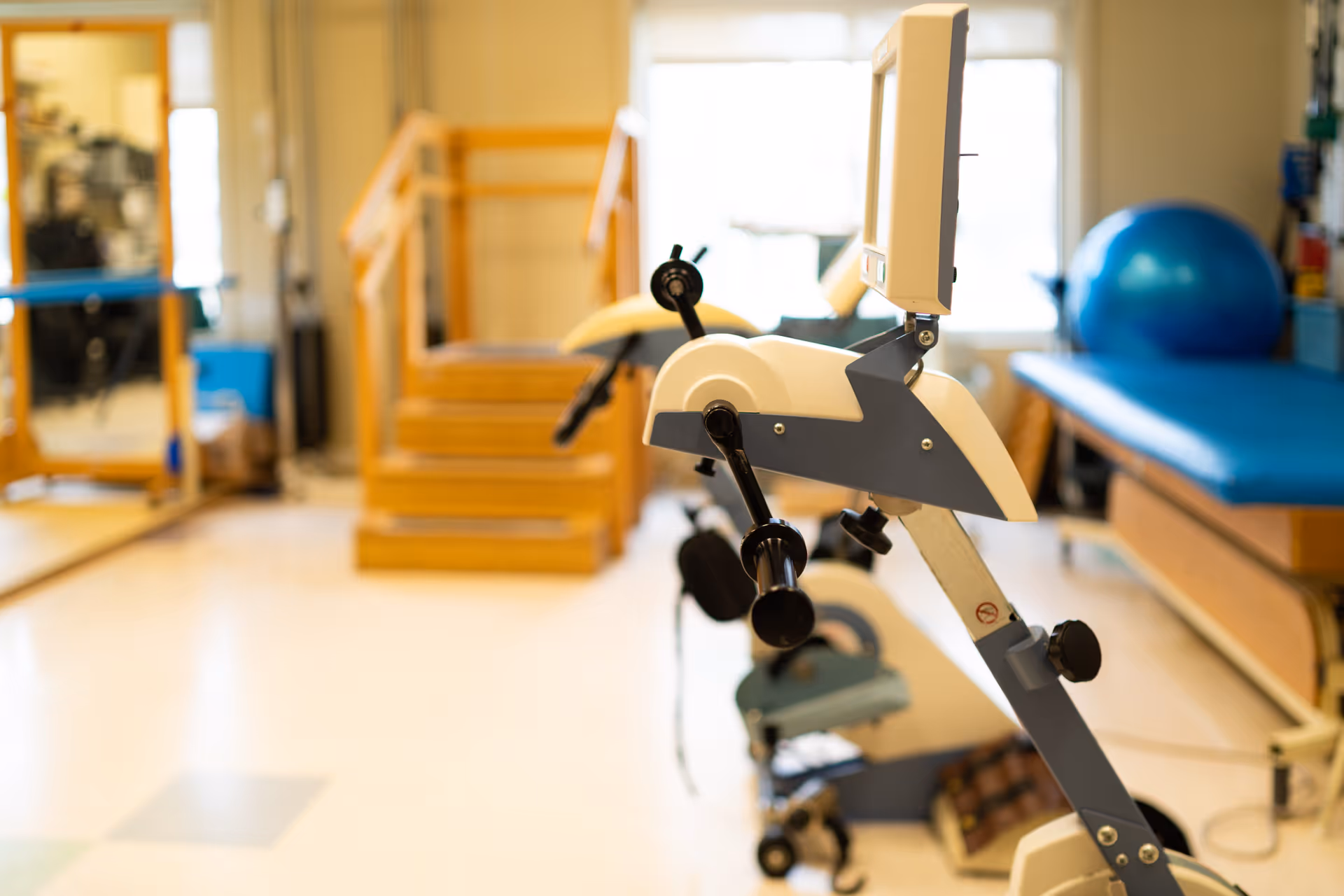 A physical therapy room with exercise equipment including a stationary bike in the foreground, wooden stairs with handrails, a blue exercise ball, and a padded therapy table near a window.
