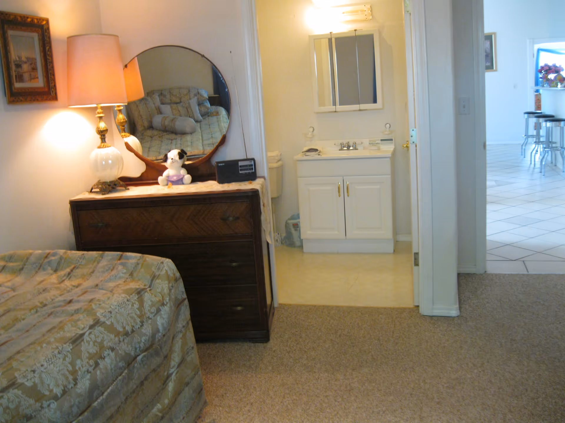 A bedroom corner with a wooden dresser topped with a round mirror, a lamp, a small stuffed dog, and a radio. The bed with a patterned bedspread is partially visible. An open doorway leads to a small bathroom with a white vanity and mirror, and beyond that, a tiled kitchen area with bar stools.