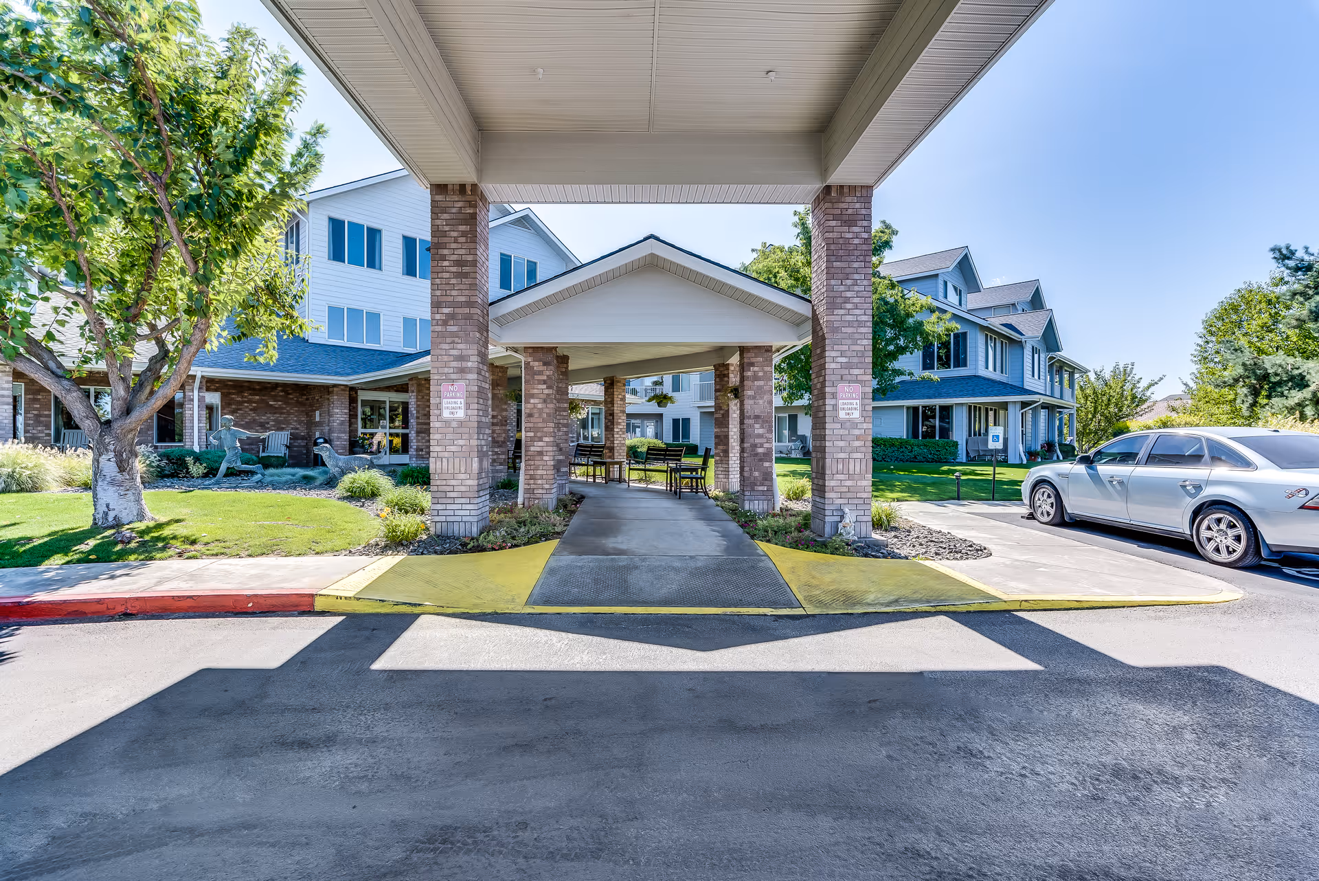 Covered porte-cochere entrance of a multi-story senior living building with brick columns, landscaped lawns, and a parked car.