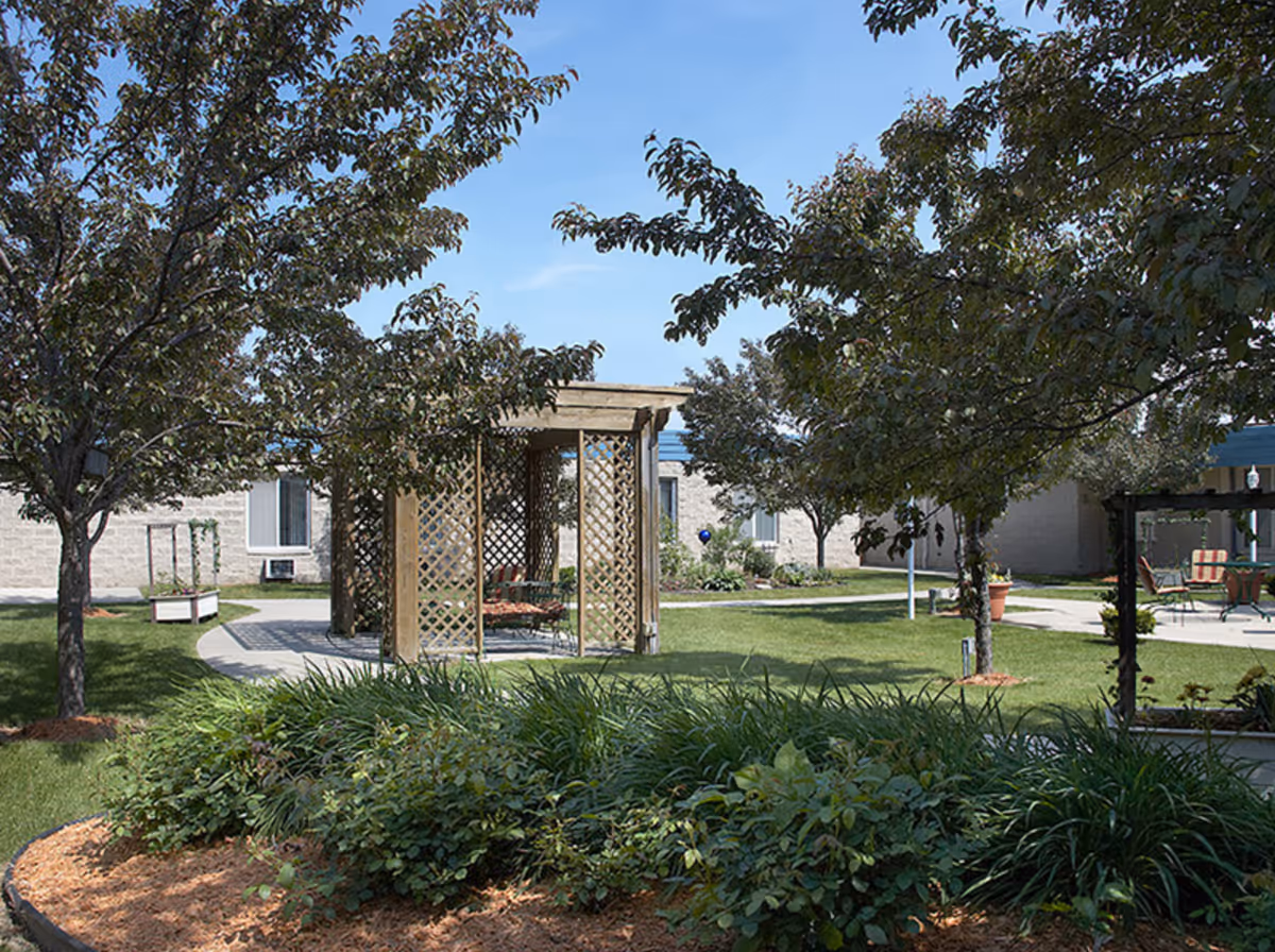 Outdoor garden area at Willowcrest Health Services featuring green grass, trees, shrubs, a wooden pergola with lattice sides, and patio furniture under a clear blue sky.