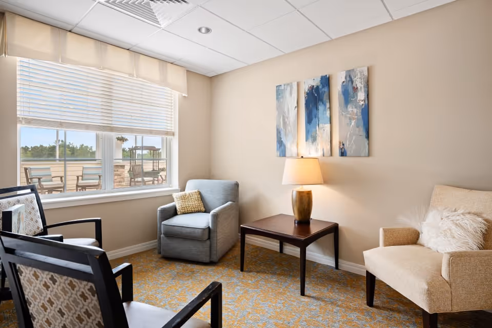 A cozy sitting area with a beige armchair with a fluffy pillow, a gray armchair with a patterned cushion, two wooden chairs with patterned upholstery, a wooden side table with a lamp, and three abstract blue and white paintings on the wall. A large window with blinds shows an outdoor patio with chairs and greenery.