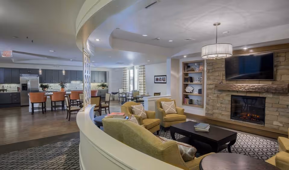 A spacious and well-lit common area featuring a cozy living room with yellow armchairs, a coffee table, a stone fireplace with a mounted TV above it, and built-in shelves. In the background, there is a kitchen area with dark cabinets, a refrigerator, and a kitchen island with orange bar stools. Several dining tables and chairs are also visible near large windows with curtains.
