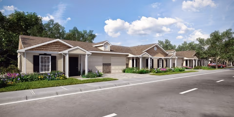 Street view of a row of single-story residential buildings with garages, small front porches, and landscaped gardens with flowers and shrubs under a partly cloudy sky.