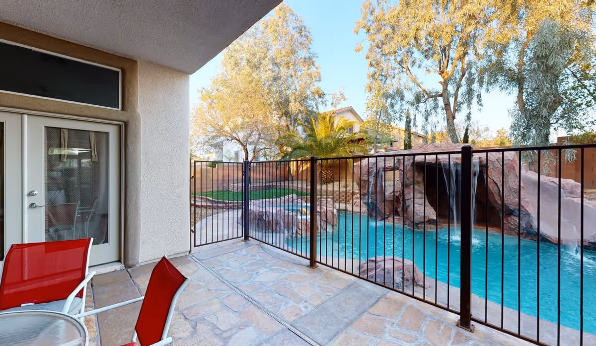 Covered patio with red chairs overlooking a fenced backyard pool featuring a rock waterfall and slide.