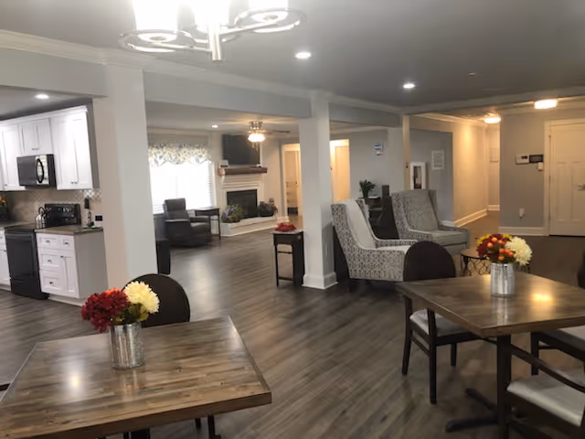Interior view of a senior living facility showing a common area with wooden tables and chairs, floral centerpieces on the tables, a seating area with armchairs, a fireplace, and a kitchen area with white cabinets and appliances in the background.