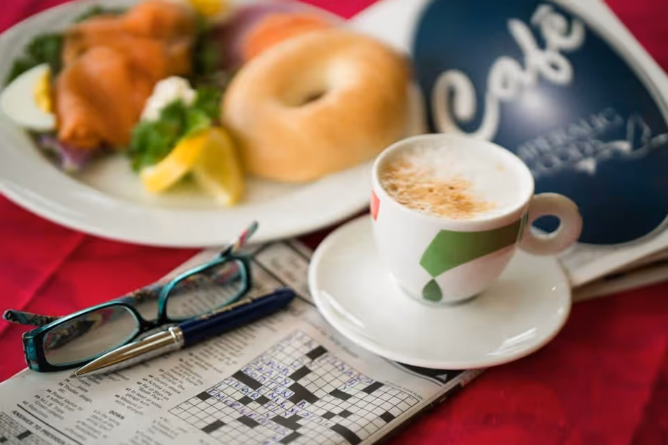 A close-up view of a breakfast setting on a red tablecloth featuring a cup of cappuccino on a white saucer, a partially completed crossword puzzle with a pen and eyeglasses resting on it, and a plate with a bagel, smoked salmon, lemon wedges, and greens. A small chalkboard sign in the background reads 'Cafe'.