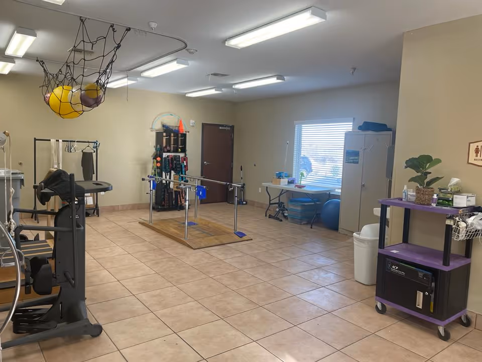 Rehabilitation gym interior with parallel bars, exercise machines, therapy balls hanging overhead, and storage carts and equipment.