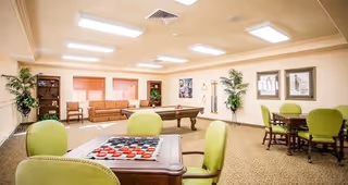 A spacious senior living common area with a pool table in the center, green cushioned chairs around tables, a checkers board on one table, a brown sofa against the far wall, potted plants, framed artwork on the walls, and bright overhead lighting.