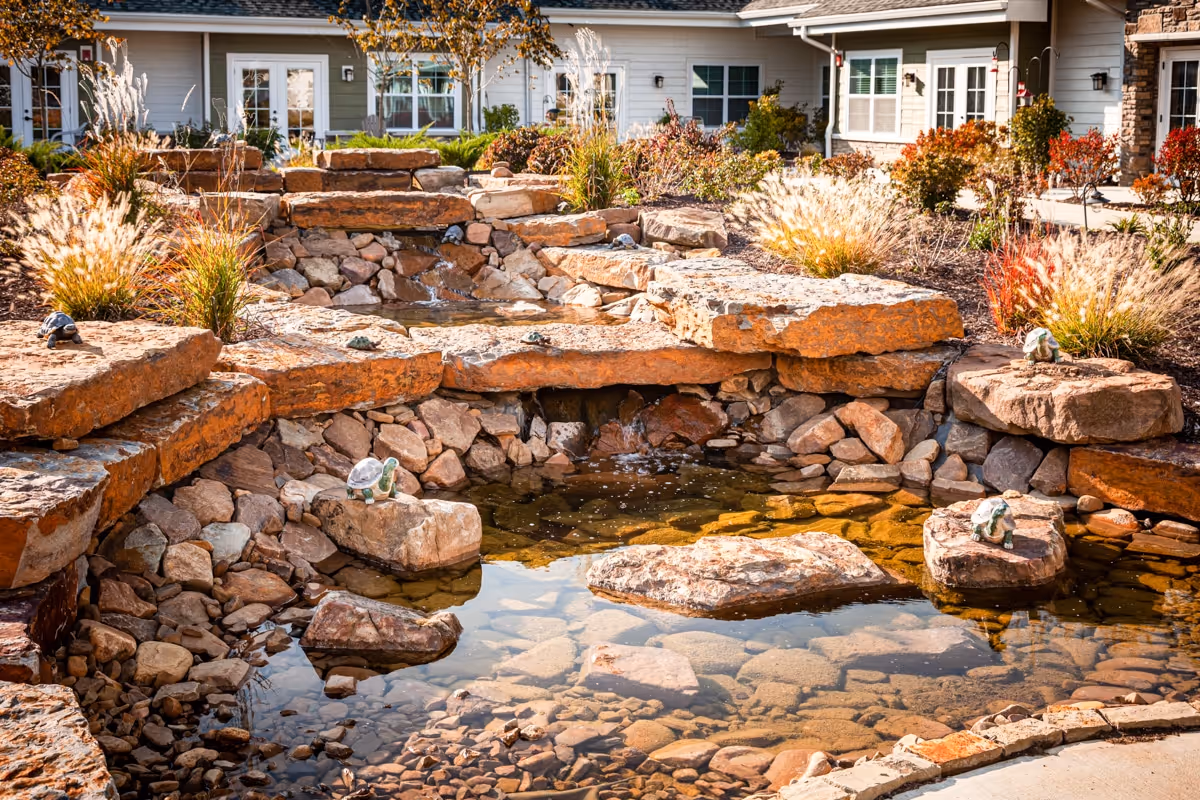 A landscaped outdoor water feature with a small pond and cascading waterfall surrounded by large rocks and ornamental grasses, with a building in the background.