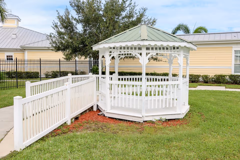White wooden gazebo with a green roof situated on a grassy lawn next to a white fence and a paved walkway, with a yellow building and trees in the background under a partly cloudy sky.
