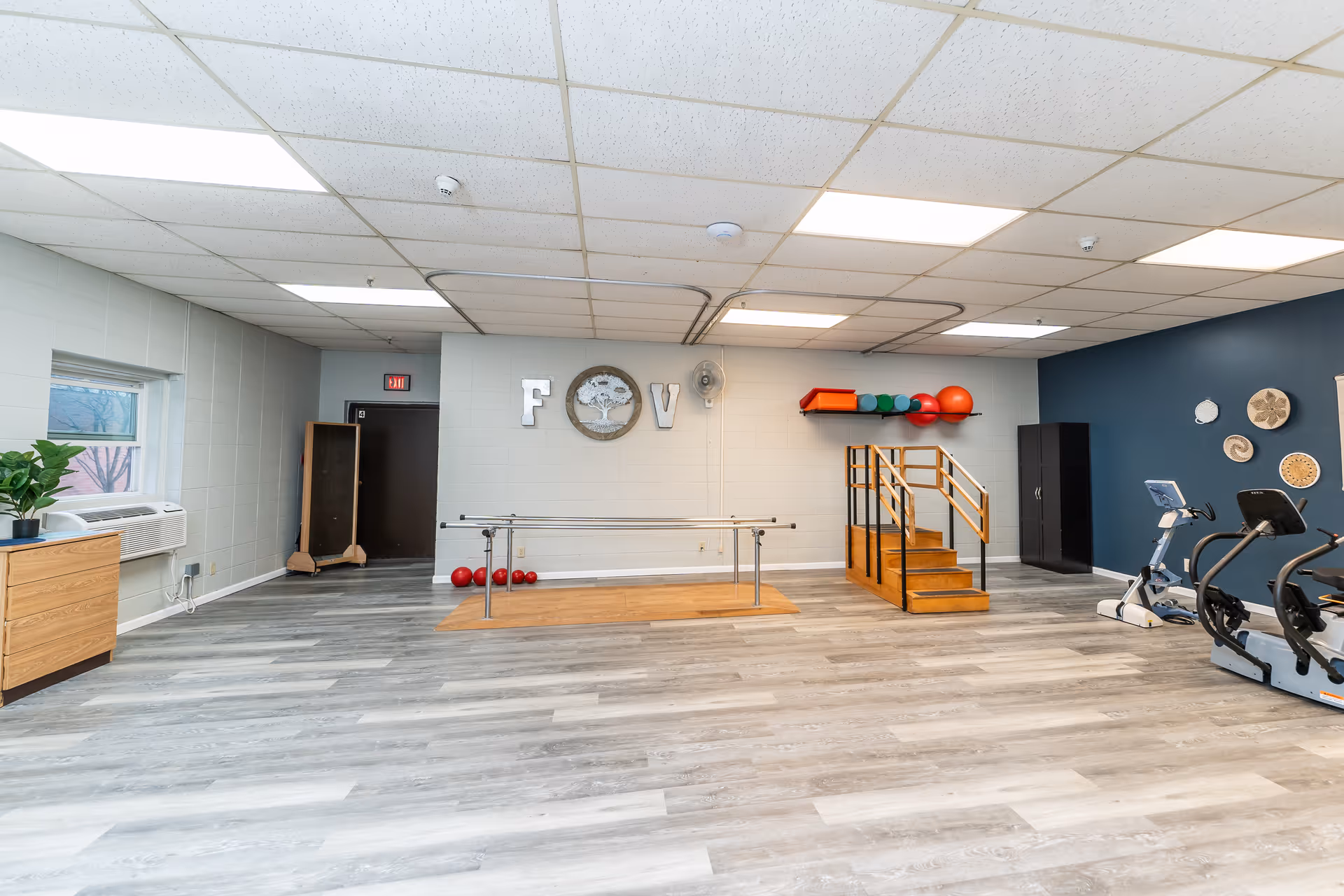 A spacious physical therapy or rehabilitation room with light gray wood flooring and white walls. The room features parallel bars for walking exercises, a small set of wooden stairs with handrails, exercise balls on a wall-mounted rack, two stationary exercise bikes, and decorative wall art. There is a window with an air conditioning unit and a wooden cabinet with a plant on top. The ceiling has fluorescent lighting panels.