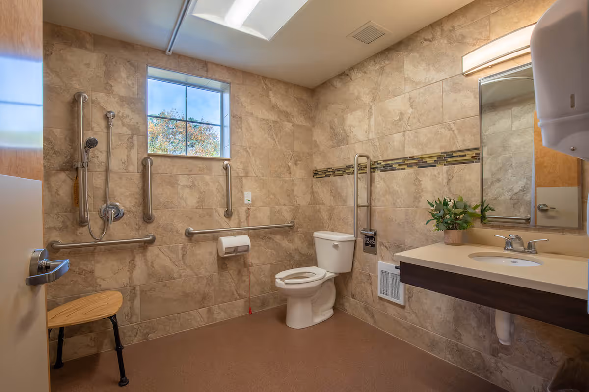 A spacious, accessible bathroom with beige tiled walls and a window. The bathroom features a toilet with grab bars, a shower area with a handheld showerhead and multiple grab bars, a wooden shower seat, a sink with a countertop, a mirror, a wall-mounted soap dispenser, and a small potted plant on the counter.