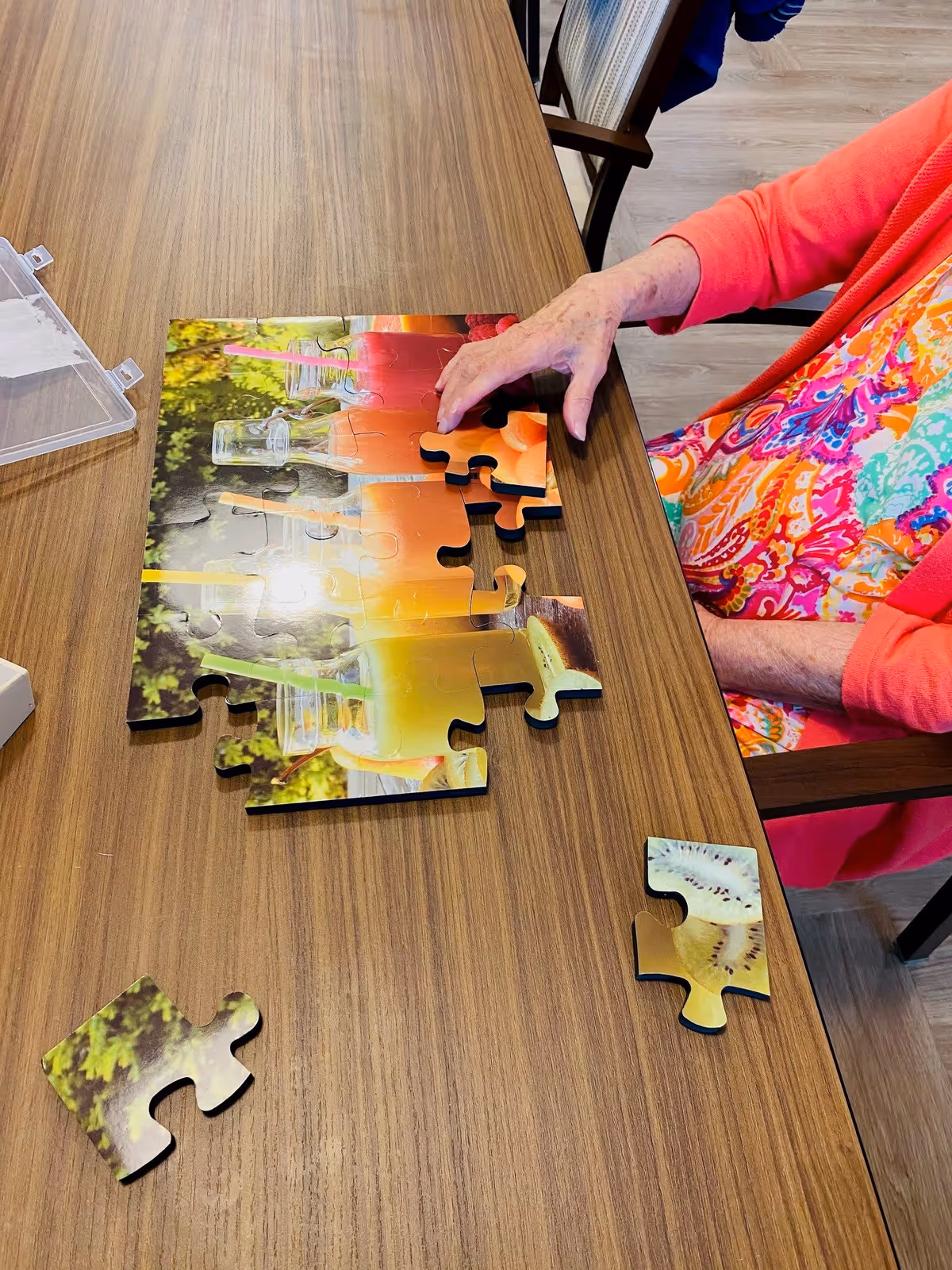 An elderly person wearing a colorful patterned shirt and coral cardigan is assembling a jigsaw puzzle on a wooden table. The puzzle features images of colorful drinks in glass bottles with straws. Two puzzle pieces are separated on the table.