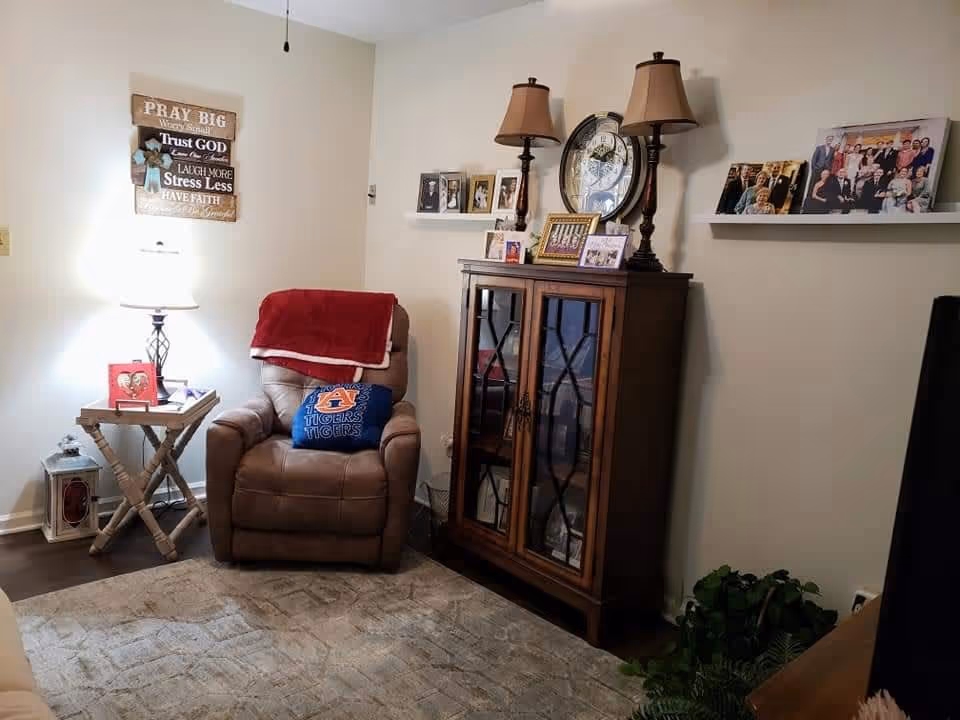 Cozy living room corner with a brown recliner chair draped with a red blanket and a blue Auburn Tigers pillow. Next to the chair is a small wooden side table with a lamp and decorative items. On the wall above the chair is a wooden sign with inspirational text. A wooden cabinet with glass doors holds framed photos and two table lamps on top. Two white floating shelves on the wall display more framed family photos. A large clock hangs on the wall behind the cabinet. The room has a beige carpet and light-colored walls.