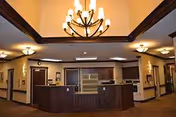 Warm-lit senior living facility lobby with curved wooden reception desk and chandeliers.