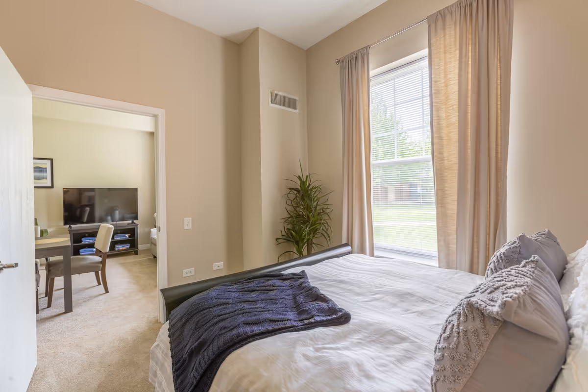 A bedroom with a neatly made bed featuring gray pillows and a dark blue throw blanket. There is a large window with beige curtains letting in natural light. A green plant is placed in the corner near the window. Through an open door, a living area with a TV, a table, and chairs is visible.