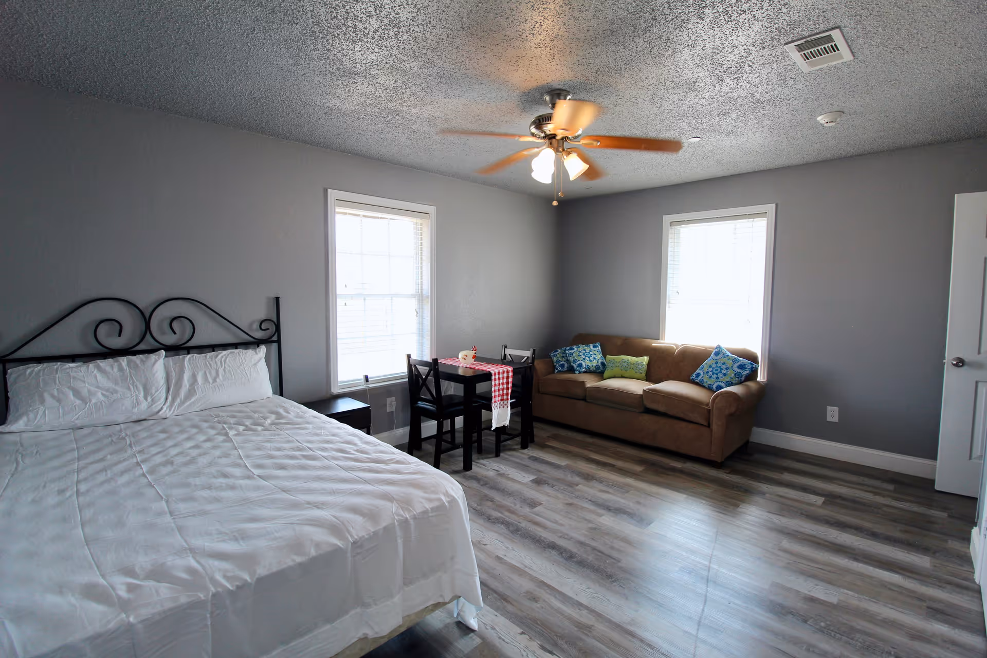 A room with gray walls and wood flooring featuring a bed with white bedding and a black metal headboard on the left, two windows with blinds, a small black table with two chairs and a red and white checkered table runner in the center, and a brown sofa with colorful pillows on the right. A ceiling fan with lights is mounted on the ceiling.