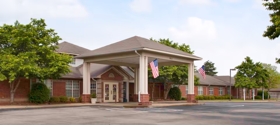 Exterior view of Wellington Manor Jackson, a single-story brick building with a covered entrance supported by columns. Two American flags hang from the entrance structure. The surrounding area has trees and a paved driveway with parking spaces.