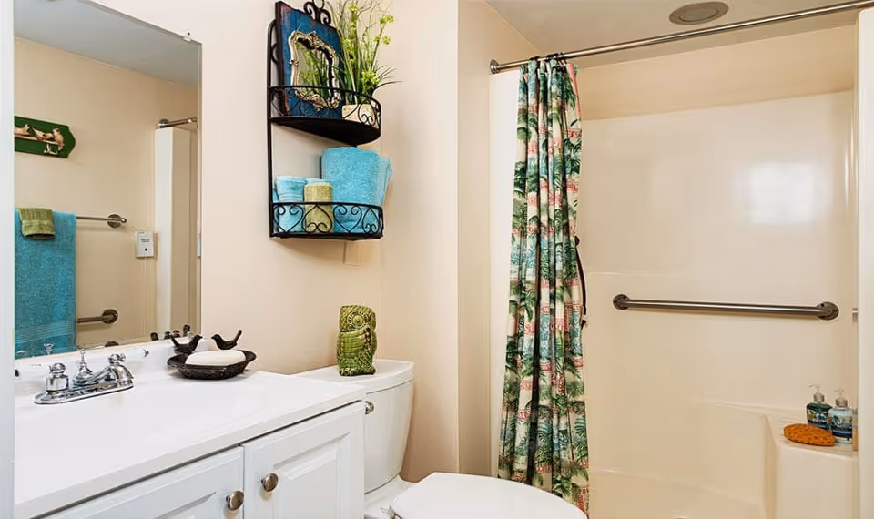 A clean bathroom featuring a white vanity with a sink and faucet, a toilet with a decorative green owl figurine on top, a wall-mounted shelf holding folded towels and a framed picture, and a shower area with a tropical-themed shower curtain and grab bar.