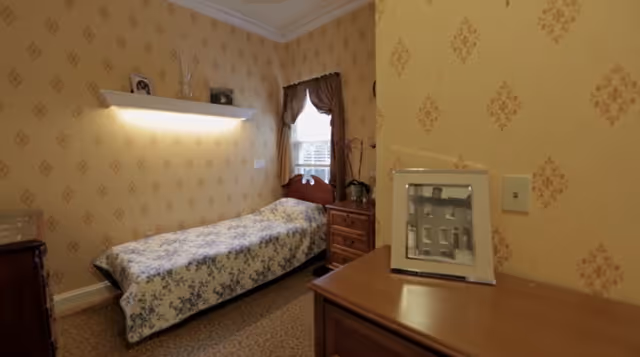 A small bedroom with a single bed covered in a white and blue floral bedspread. The room has patterned wallpaper in a beige and gold color scheme. There is a wooden nightstand with a plant and a window with brown curtains. A floating shelf above the bed holds framed photos and decorative items. A wooden dresser with a framed black and white photo is visible in the foreground.