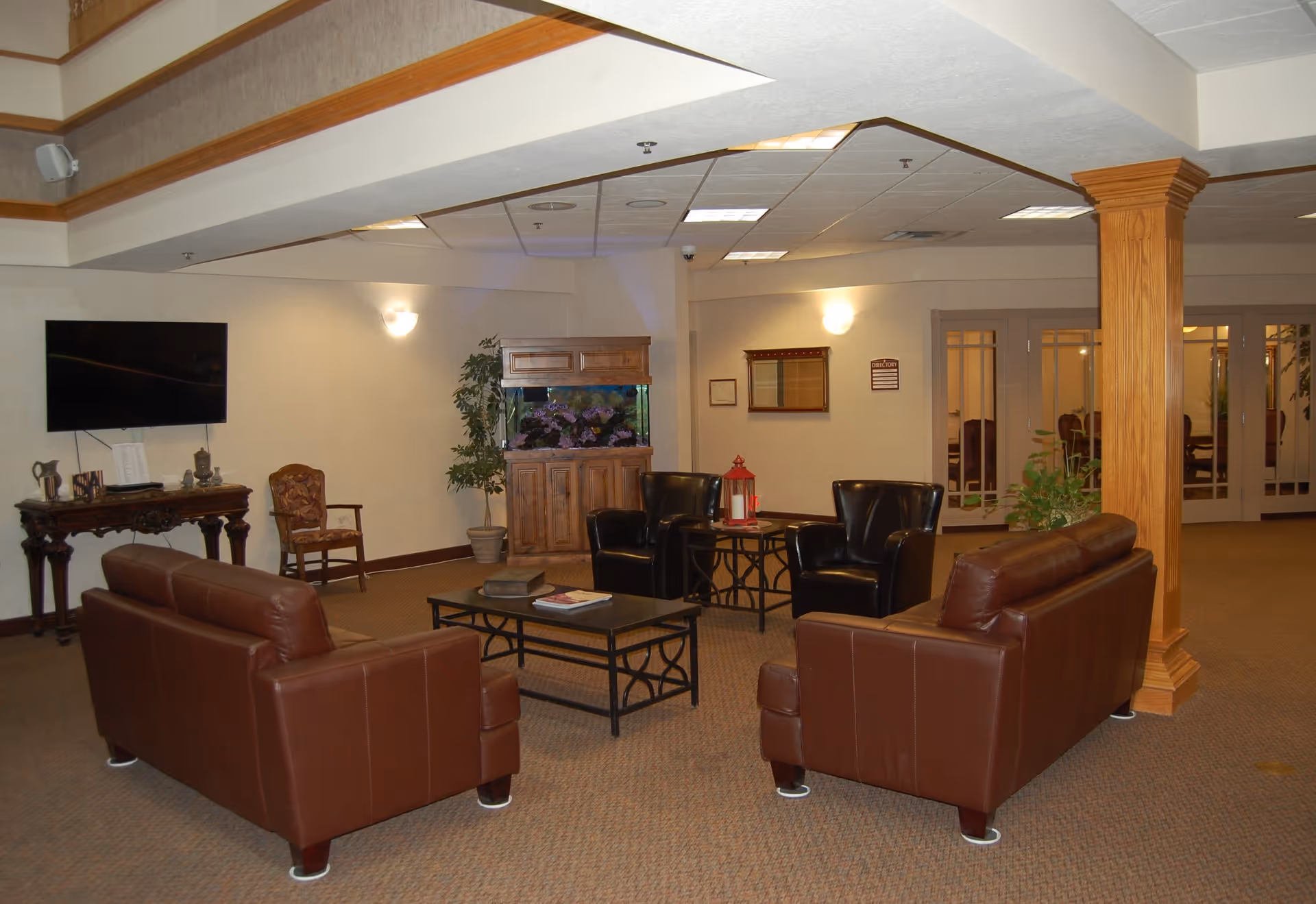 A cozy living room area with two brown leather sofas facing each other, two black leather armchairs, a coffee table with books, and a side table with a red lantern. There is a large fish tank on a wooden cabinet, a flat-screen TV mounted on the wall, a wooden chair, and a decorative wooden table. The room has beige walls, carpeted floor, and a wooden column supporting the ceiling.