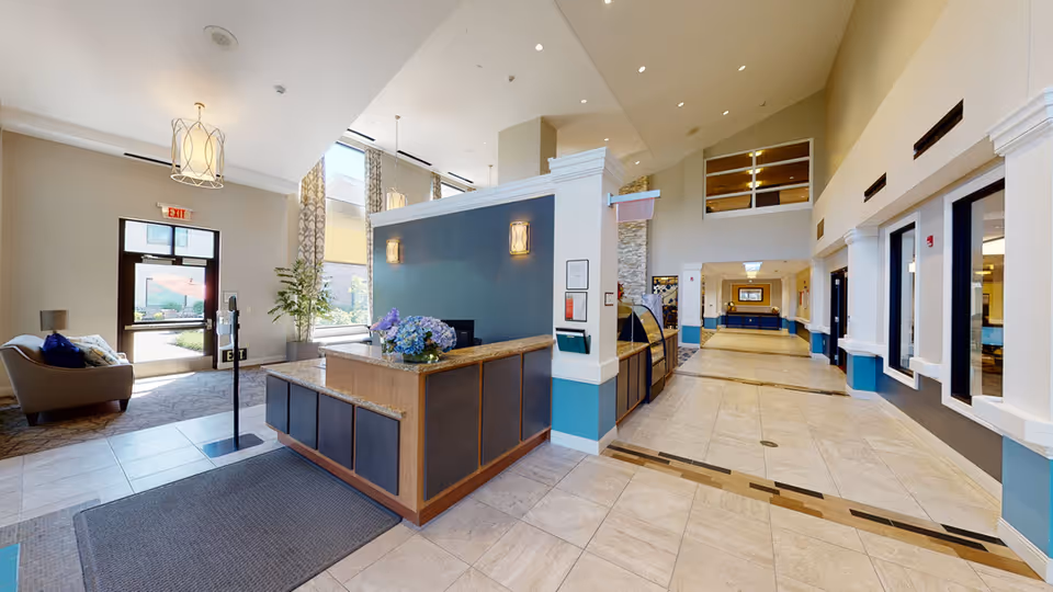 Bright and spacious reception area of ProMedica Total Rehab + (Exton) with a front desk featuring a granite countertop and a flower arrangement. The area includes large windows allowing natural light, a seating area with a couch and pillows near the entrance door, decorative hanging lights, and a long hallway leading further into the facility.