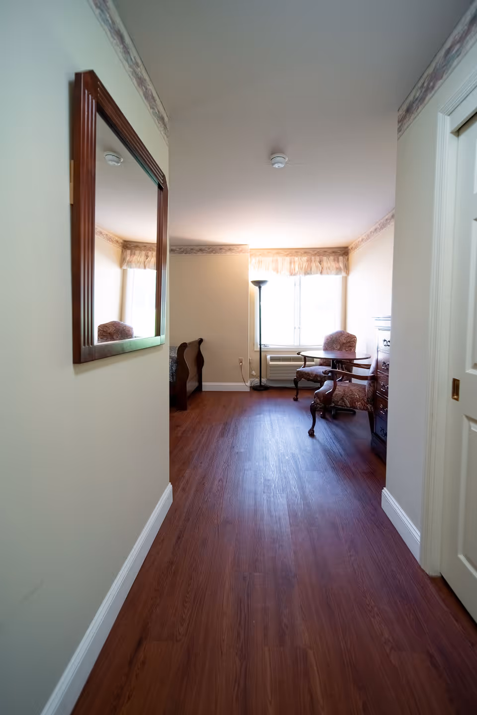 View down a hallway with wooden flooring leading to a room with a window. The room contains a wooden table with two upholstered chairs, a floor lamp, and a wooden dresser. A large mirror is mounted on the left wall of the hallway.