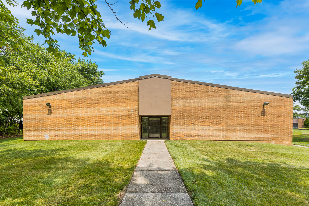 Exterior view of a single-story brick building with a central entrance featuring glass doors. The building is surrounded by green grass and trees under a partly cloudy blue sky.