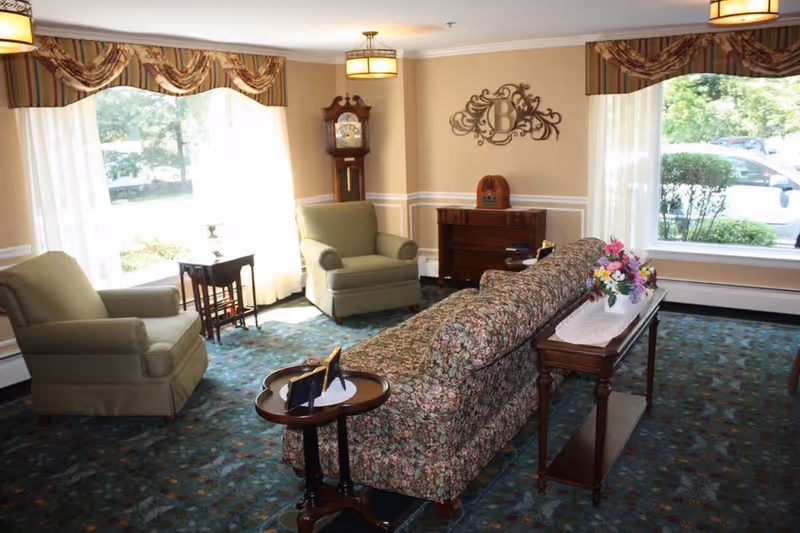 A cozy living room with two green armchairs, a floral patterned sofa, a wooden side table with a flower arrangement, a grandfather clock, and large windows with sheer white curtains and decorative valances letting in natural light.