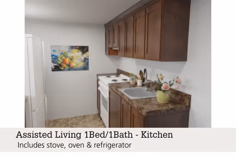 A small kitchen area with dark wooden cabinets, a white stove and oven, a white refrigerator, and a countertop with a sink and a small flower pot. There is a colorful painting on the wall.