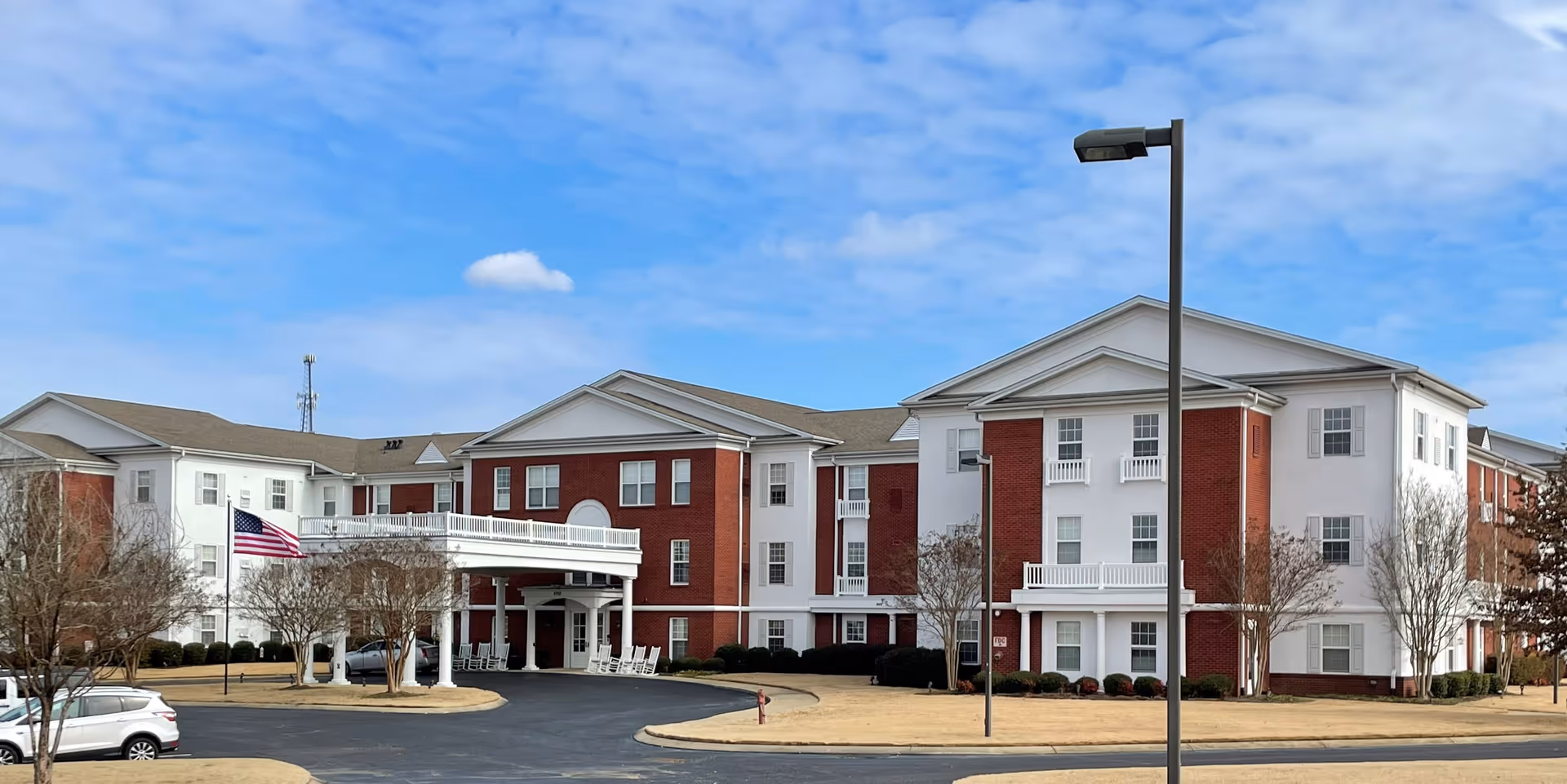 Exterior view of The Goldton at Southaven, a large three-story senior living facility with red brick and white siding. The building features a covered entrance with white columns, rocking chairs, and an American flag on a flagpole. The sky is partly cloudy and the surrounding area has trimmed bushes and leafless trees.