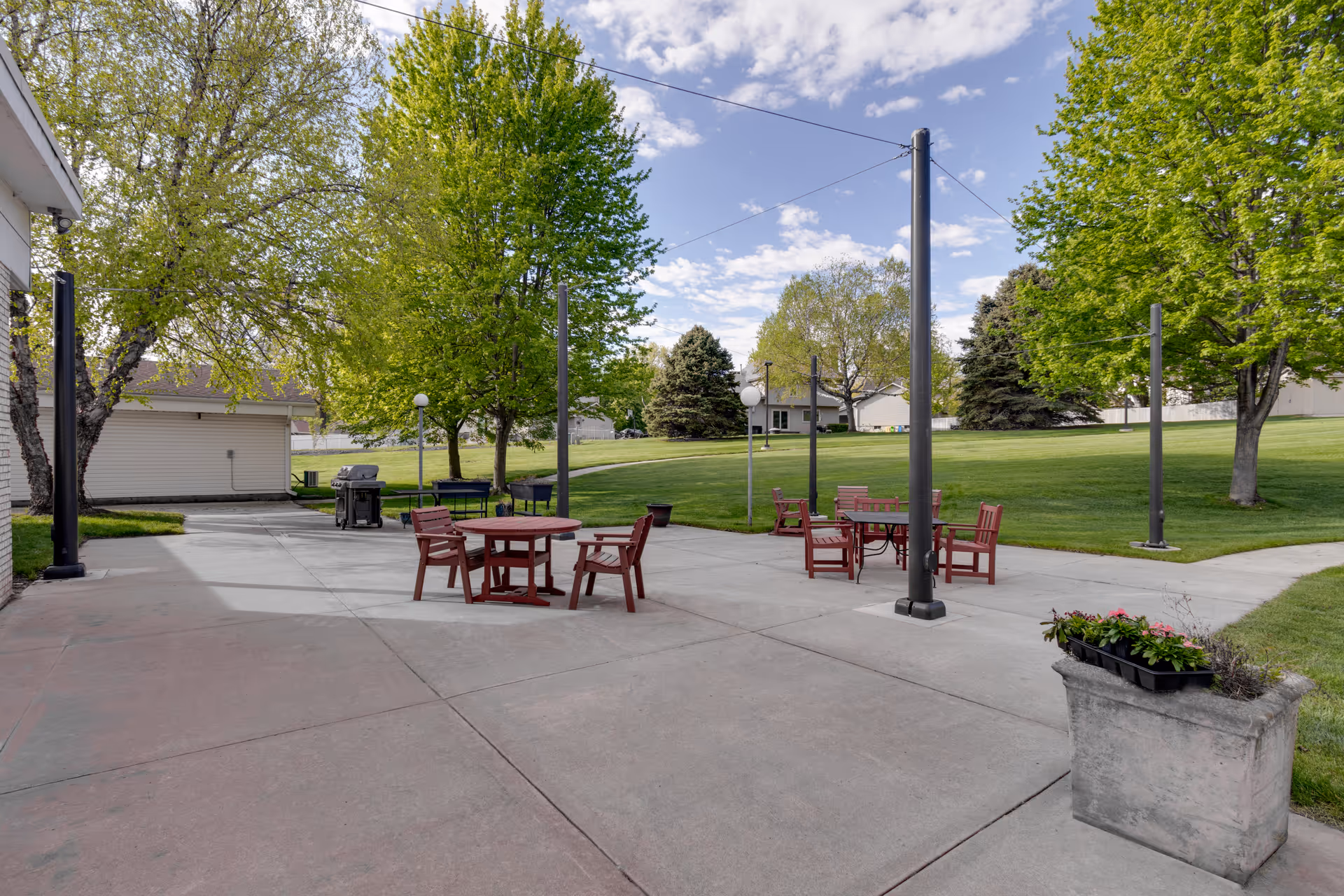 Outdoor patio area with several round wooden tables and chairs on a concrete surface, surrounded by green grass and trees under a partly cloudy sky. There are light poles and a barbecue grill visible near the building on the left side.