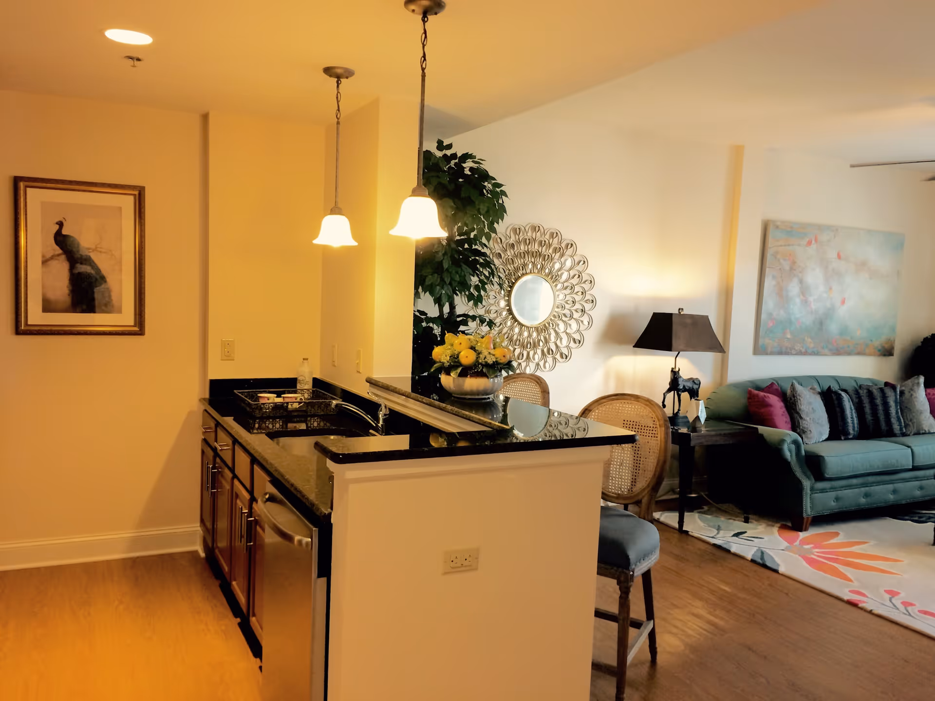 Interior view of a senior living facility showing a kitchen area with dark granite countertops, wooden cabinets, and two pendant lights hanging above the counter. Adjacent to the kitchen is a living room with a green sofa, decorative pillows, a side table with a lamp, a round decorative mirror on the wall, and a colorful area rug on the wooden floor.