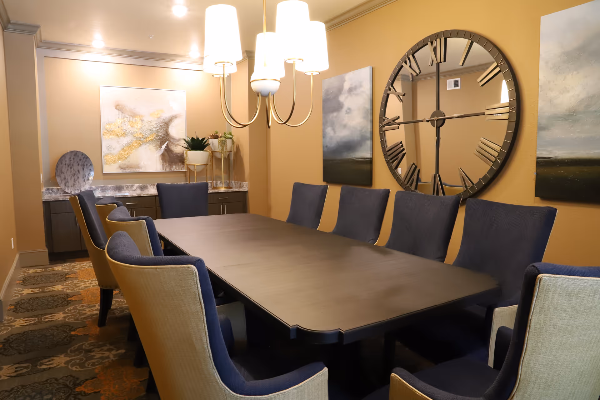 A dining room with a long dark wooden table surrounded by eight upholstered chairs in navy blue and beige. The walls are painted a warm beige color and decorated with two landscape paintings and a large round wall clock with Roman numerals. A modern chandelier with five lights hangs above the table. There is a sideboard with decorative items and plants in the background.