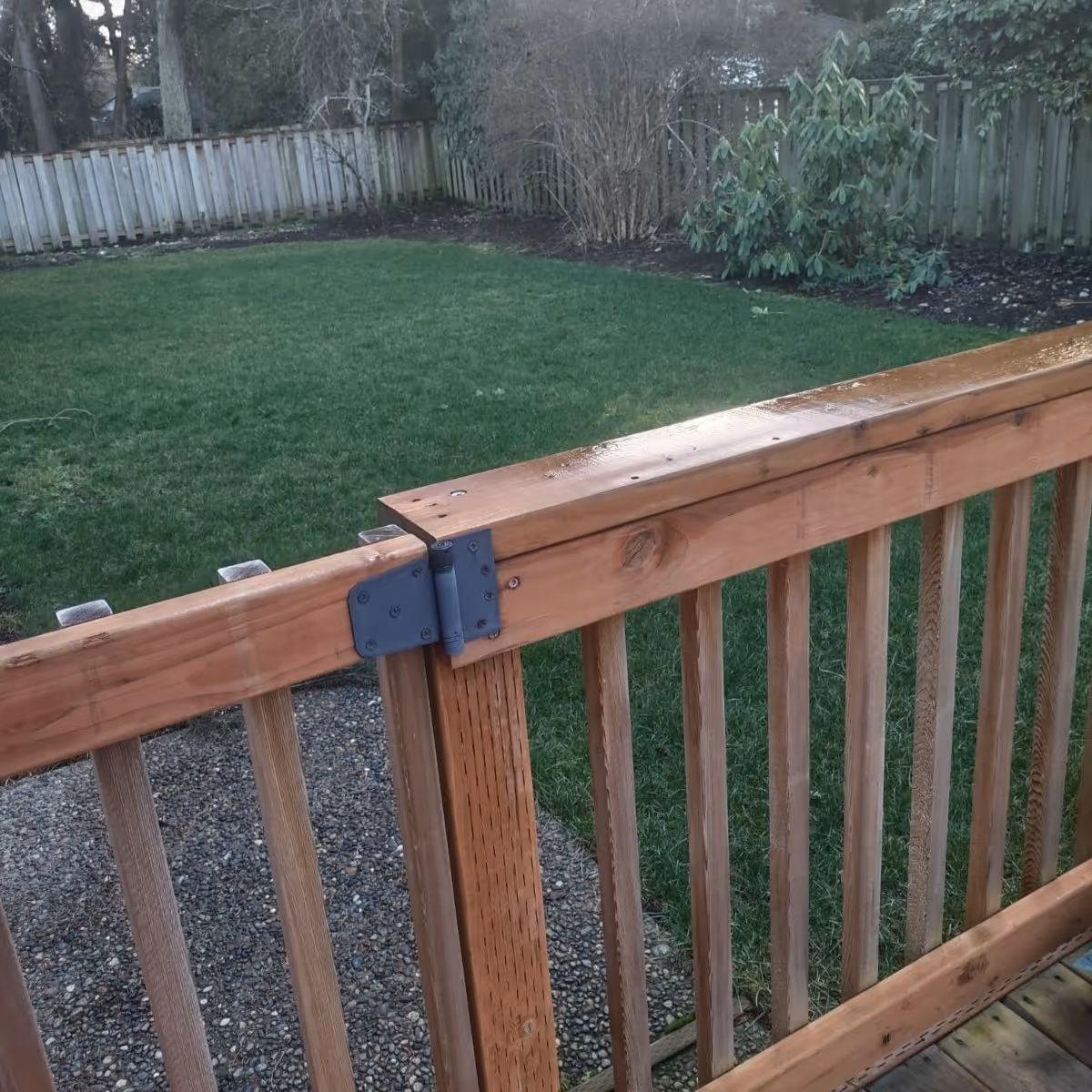 View of a backyard with a green lawn, bordered by a wooden fence and some bushes. In the foreground, there is a wooden railing with a metal hinge, possibly part of a gate or deck.