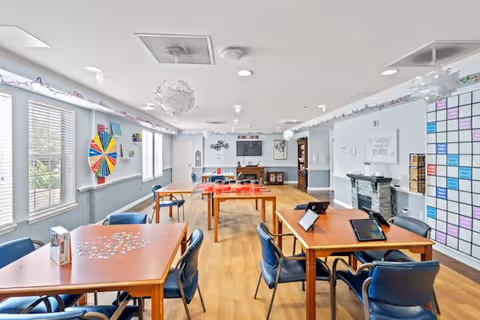 Bright communal activity room with wooden tables and blue chairs, puzzles and tablets on the tables, a colorful prize wheel by the windows, and a TV on the far wall.