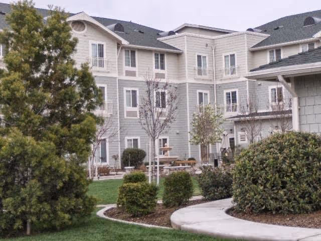 Exterior view of a multi-story senior living facility building with light gray siding and white trim. The foreground features a landscaped garden area with green bushes, trees, and a curved concrete walkway.