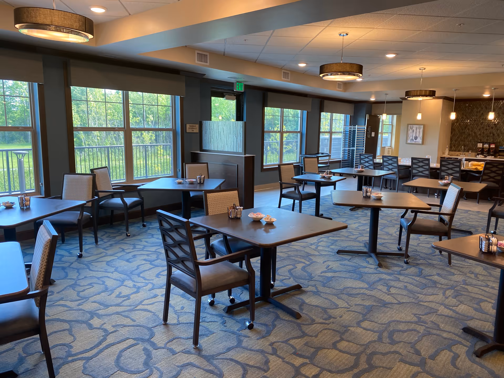 A spacious dining room with multiple square tables and chairs arranged neatly. Large windows on one side allow natural light to fill the room, showing a view of green trees outside. The room has patterned carpet flooring, modern ceiling lights, and a counter area with coffee machines and other items in the background.