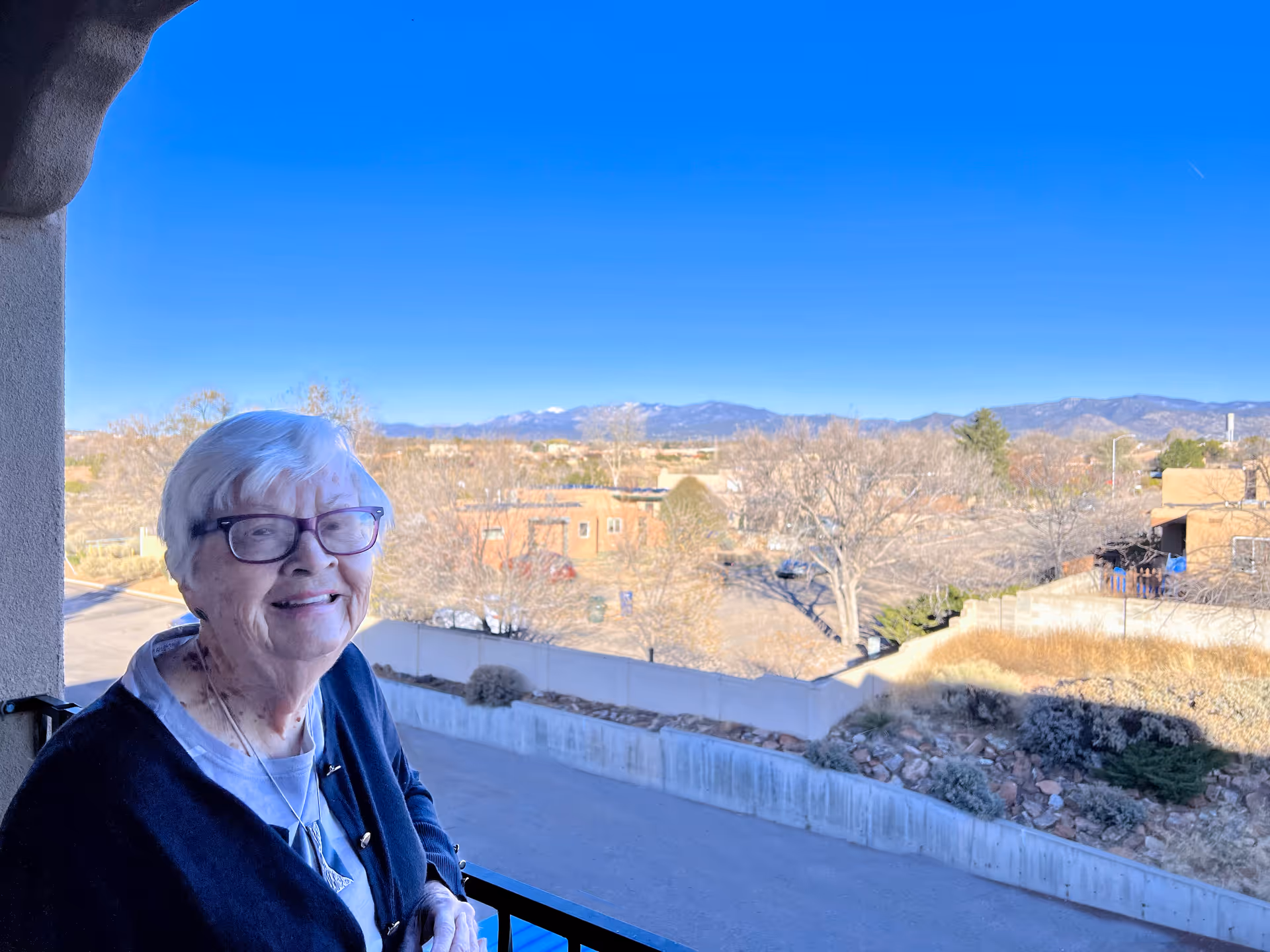An elderly woman with white hair and glasses smiling while standing on a balcony overlooking a suburban area with trees, houses, and mountains in the background under a clear blue sky.