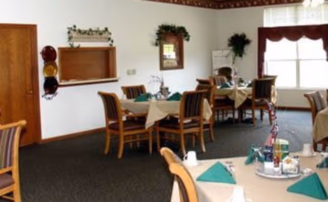 A dining room with several tables covered with beige tablecloths and green napkins. Each table is surrounded by wooden chairs with striped cushions. The room has a large window with a valance, a wooden door, and a wall-mounted mirror with greenery above it. There are decorative plants and a serving pass-through window on one wall.
