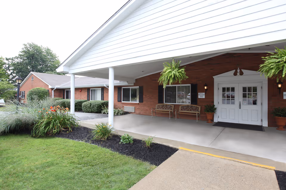 Entrance to a brick building with a white covered porch supported by white columns. There are two benches and potted plants near the double white doors. The surrounding area has green grass, bushes, and flowering plants.