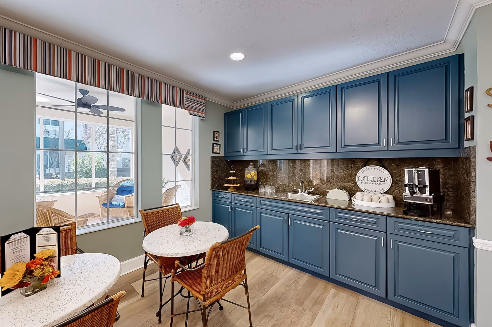 A cozy kitchen area with blue cabinets and a granite countertop featuring a coffee bar setup. There are two small round tables with wicker chairs, each table decorated with a small vase of flowers. Large windows with striped valances let in natural light and offer a view of an outdoor seating area with wicker furniture and a ceiling fan.