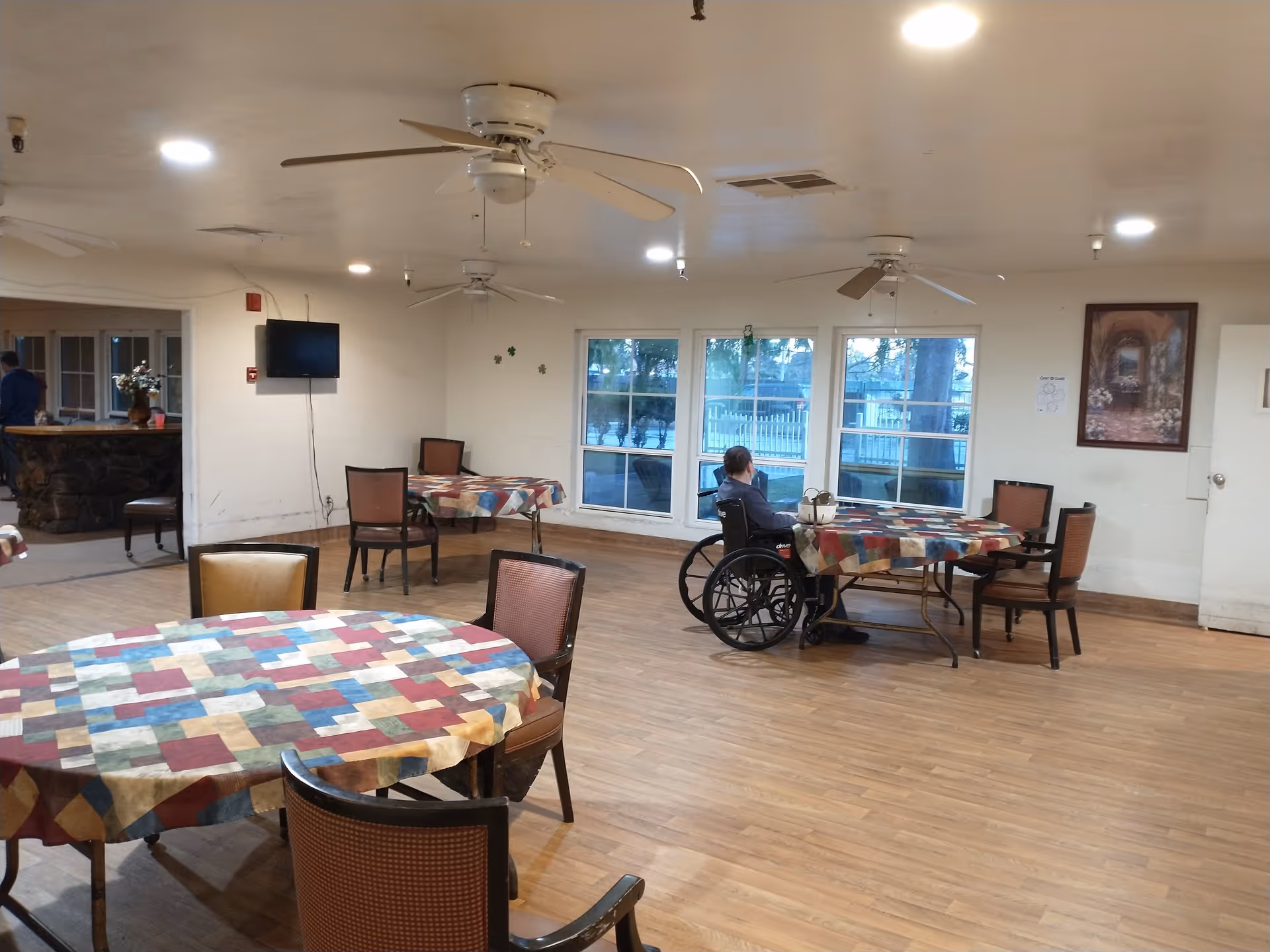 A dining room in an assisted living facility with several round tables covered with colorful patchwork tablecloths. There are chairs around the tables, and one person in a wheelchair is seated at a table near large windows. The room has ceiling fans, recessed lighting, and a wall-mounted TV. A framed picture and some decorations are on the walls.