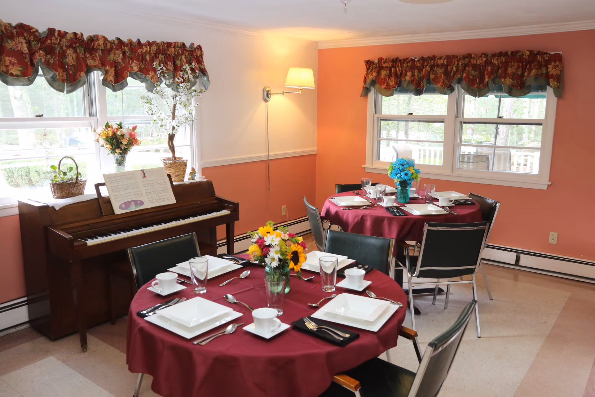 A dining room with two round tables covered in burgundy tablecloths, each set with white square plates, cups, glasses, and silverware. Each table has a vase with colorful flowers. There is a piano against the wall with sheet music on it, and windows with floral valances letting in natural light. The walls are painted peach and white, and there is a wall-mounted lamp providing additional light.
