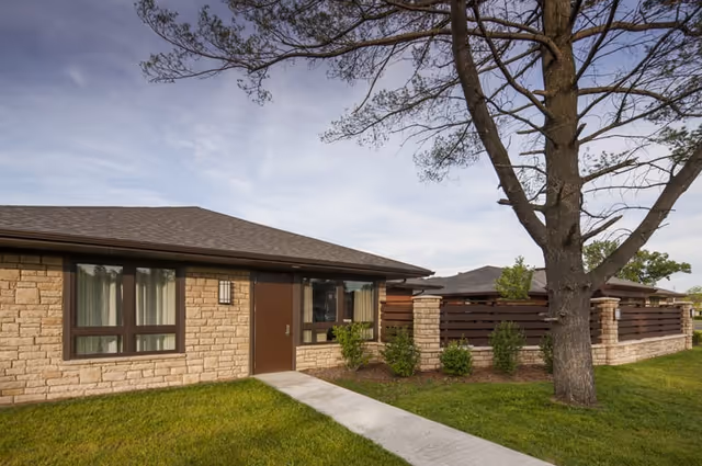 Single-story stone-faced building entrance with a sidewalk, lawn, large tree, and wooden privacy fence.