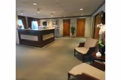 Interior view of a nursing facility reception area with a front desk, chairs, a small table with a plant, and doors leading to other rooms. The space is carpeted and well-lit with ceiling lights and windows with curtains.
