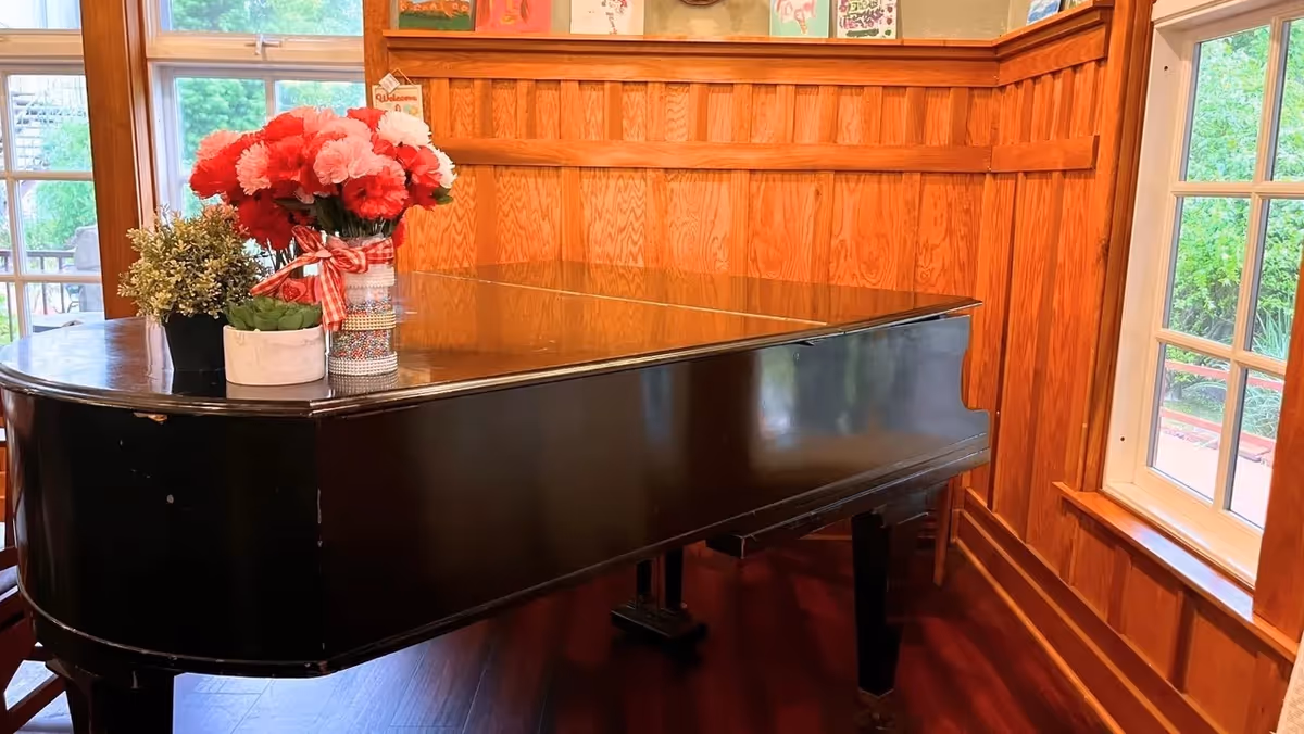A black grand piano in a corner of a room with wooden panel walls and large windows showing greenery outside. On top of the piano are three flower arrangements, including red and white flowers in a vase with a red and white checkered ribbon.