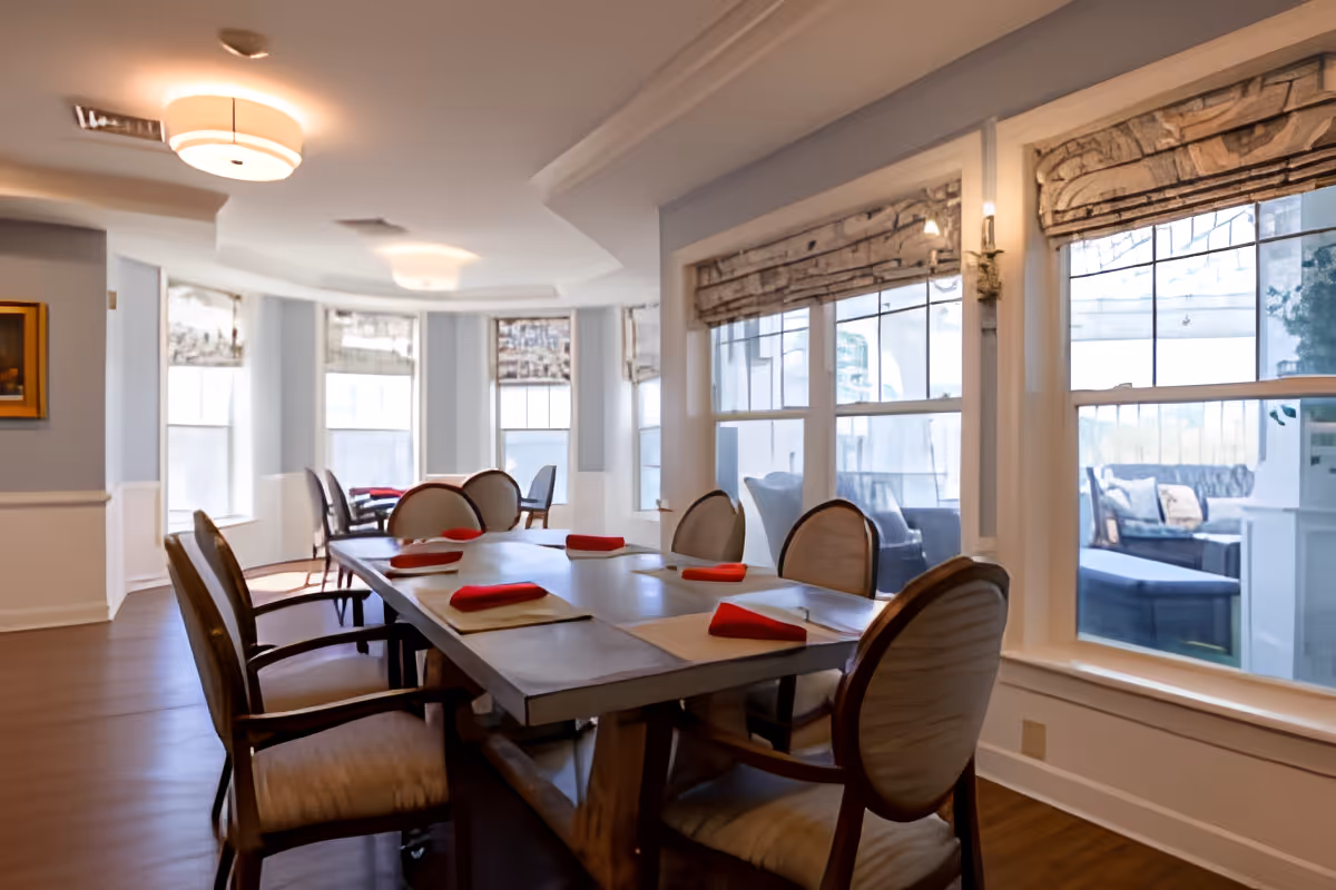 A bright dining room with a large rectangular table set with placemats and red folded napkins. The room features several upholstered chairs around the table, large windows with patterned valances, and soft ceiling lights. Outside the windows, a patio area with outdoor seating is visible.