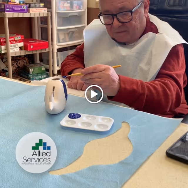 An elderly man wearing glasses and a white bib is painting a small ceramic object with blue paint at a table covered with a blue cloth. There is a paint palette with blue paint on the table, and shelves with board games in the background.